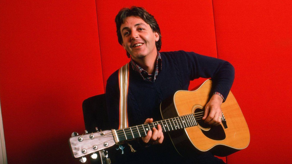 Portrait of British musician Paul McCartney as he plays acoustic guitar against a red background, October 7, 1984 . Crédits : Robert R. McElroy