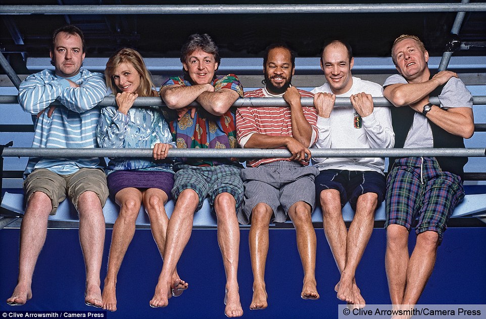 A laid back shot of Paul and Linda McCartney and his touring band on the cover of Off The Ground sitting with their trousers rolled up and their feet dangling off the edge of a railing