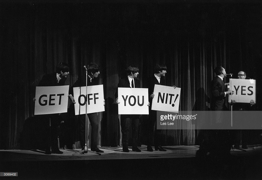 The Beatles on stage at the London Palladium hold placards saying,'Get Off You Nit'.