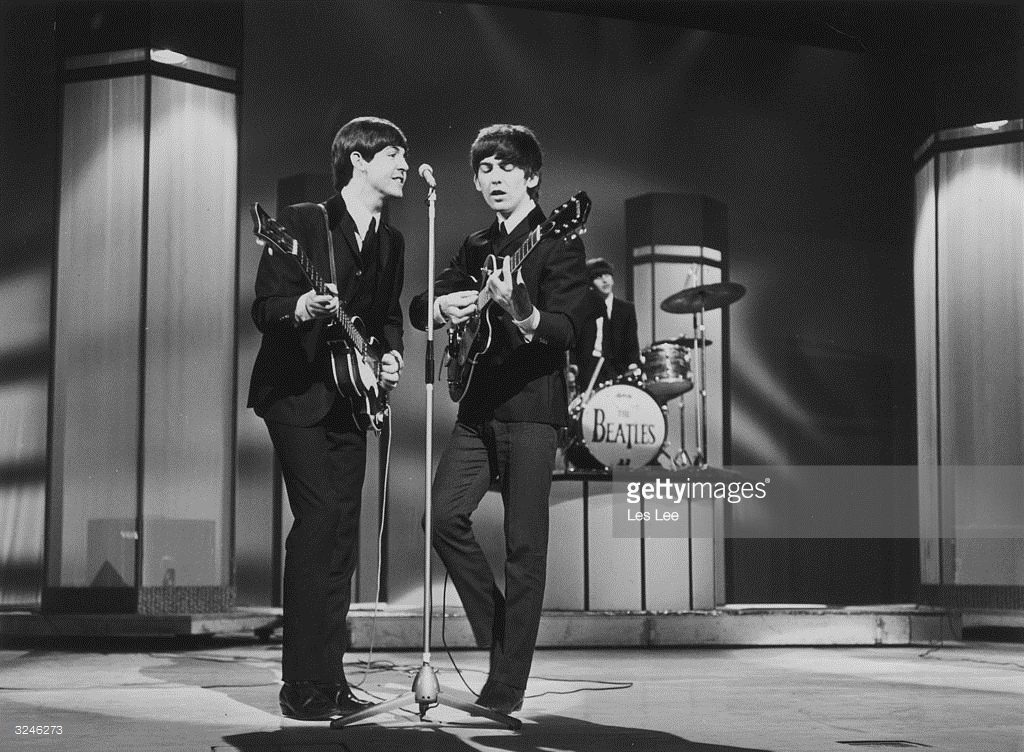 Bassist Paul McCartney and guitarist George Harrison (1943 - 2001) sing at a microphone as the Beatles perform at the London Palladium. Drummer Ringo Starr plays from a riser in the background. London, England, January 1964.